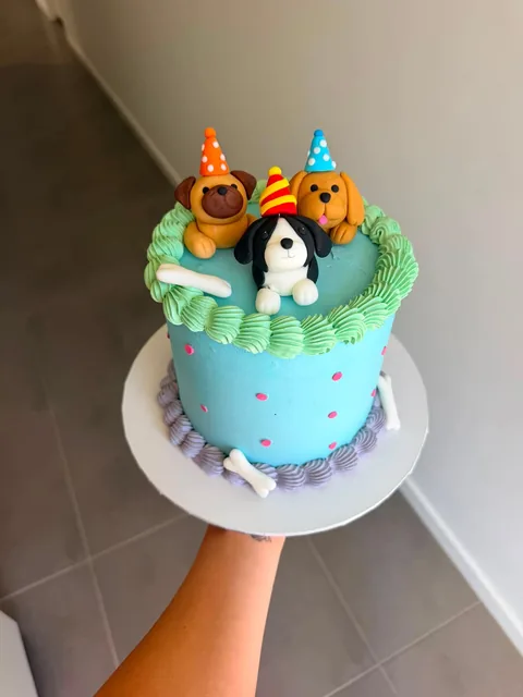 Tall blue buttercream cake with a green piped border, pink dots, fondant bones and three fondant puppies in party hats